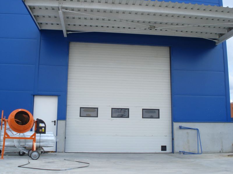 Ușă de garaj industrială albă cu hublouri, montată la o hală de producție