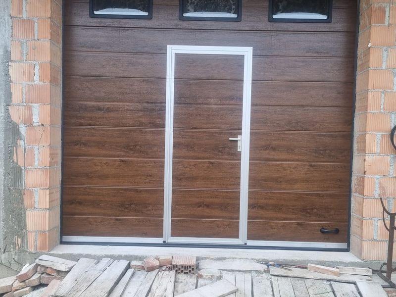 Ușă de garaj secțională nuc cu ușă pietonală albă și hublouri, la o casă în construcție
